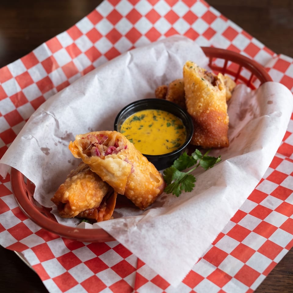 Golden-brown egg rolls with a side of yellow dipping sauce and cilantro in a red basket.