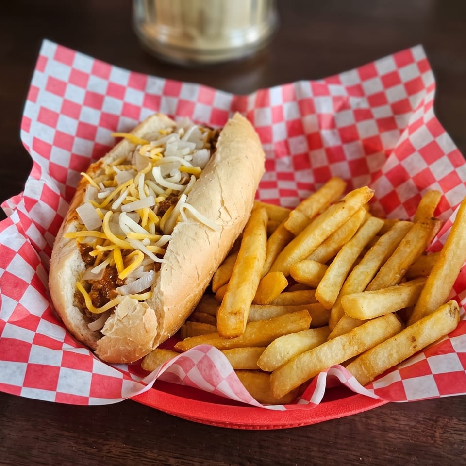 Chili dog with shredded cheese and diced onions, served with golden french fries in a red basket.
